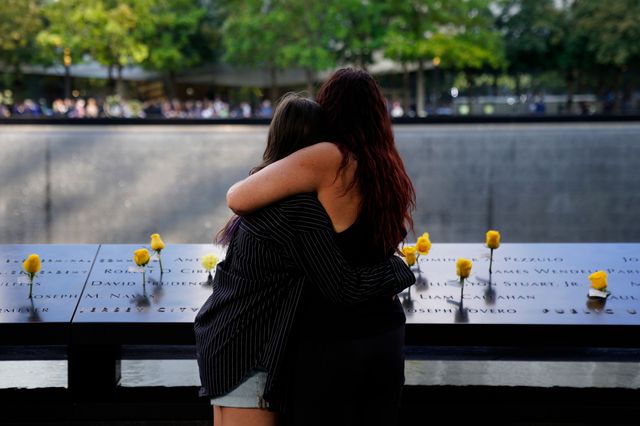 Two people embrace each other while looking at the names inscribed in the 9/11 memorial, which has yellow roses inserts in some names